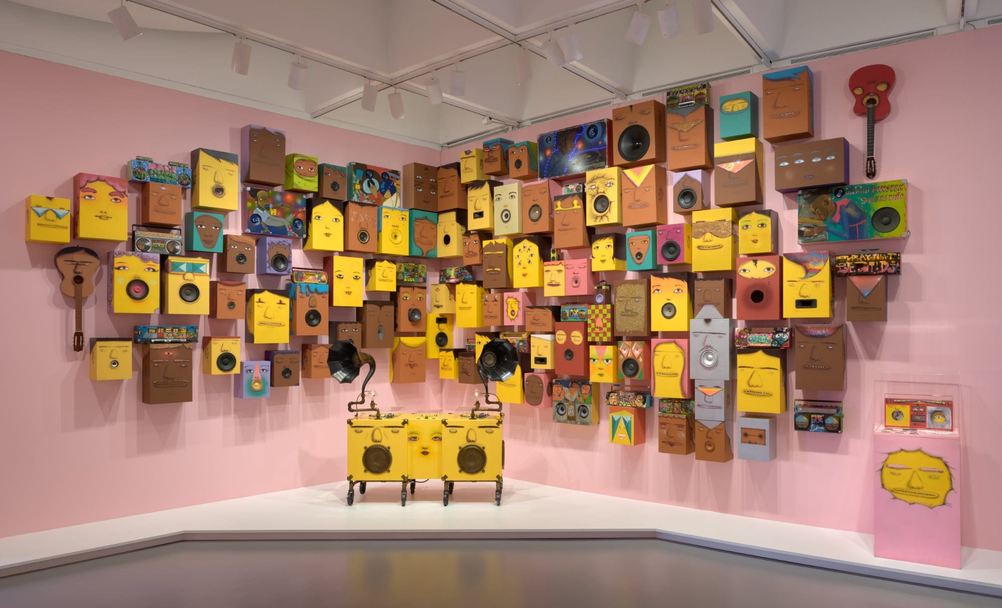 a collection of speakers painted with yellow and brown faces against a pink wall. a gramophone and boombox painted similarly stand in the front