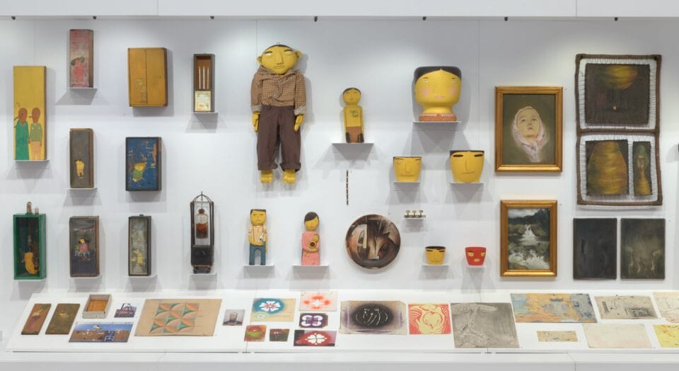 a collection of drawings, paintings, and figurative sculptures in yellow hang in a display case