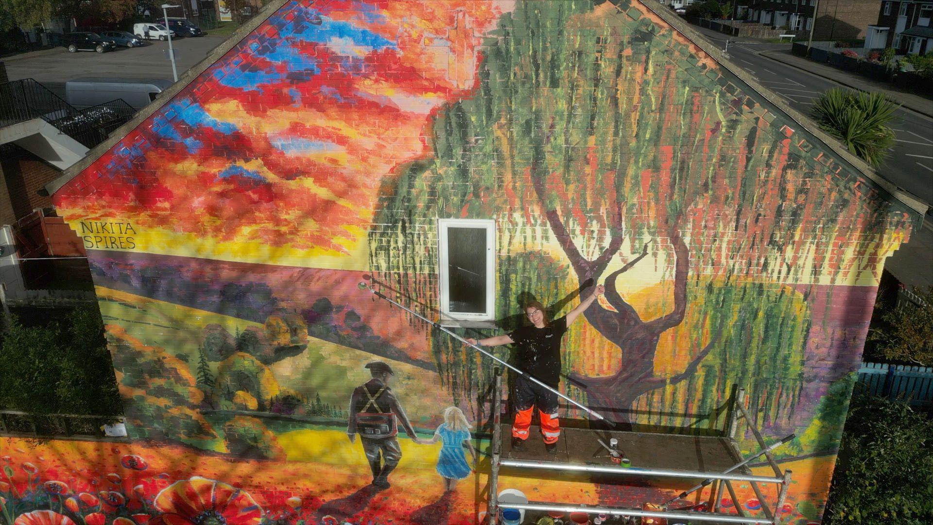 Nikita stands on scaffolding in front of the mural holding a roller with her right hand. She has her arms outstretched and is smiling. She is wearing glasses, fluorescent orange work trousers and a black t-shirt. The mural: in the foreground we can see red poppies with fields stretching out behind them as the sun sets. A soldier walks away holding hands with his daughter. He is dressed in a military uniform and his daughter has blonde hair and a light blue dress. Above them there is a large willow tree.