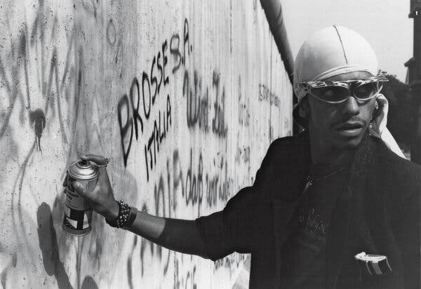 A black-and-white photo of the artist Rammellzee spray-painting a concrete wall with graffiti.