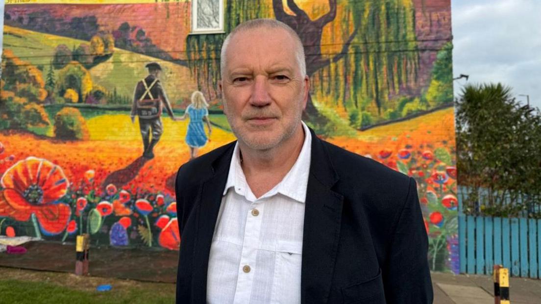 Steve Holland stands in front of the mural looking down the lens. He has gray hair and is wearing an open-collared cream linen shirt with a black blazer.