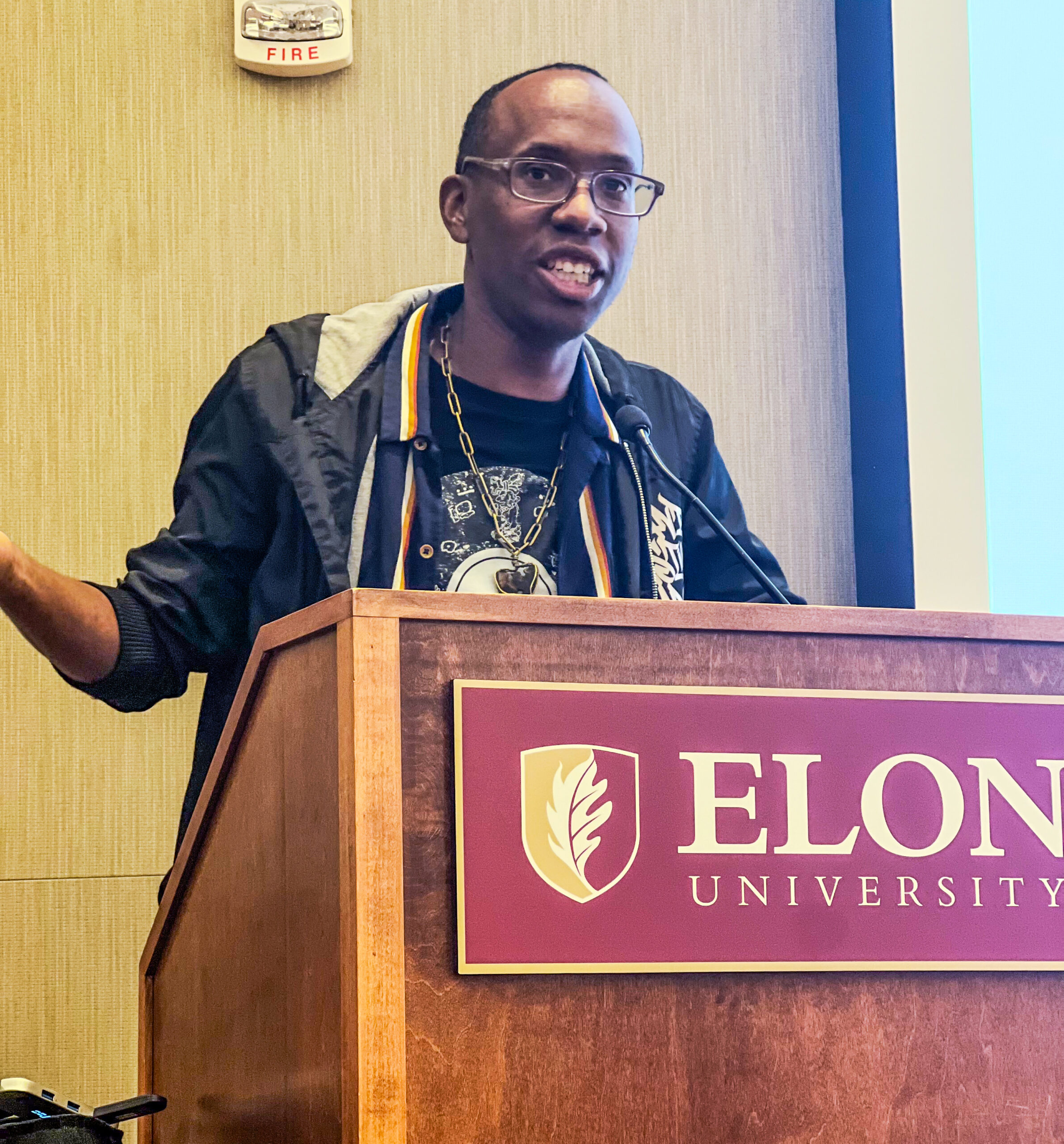 A man speaking at a podium with Elon University's signature in McBride Gathering Space