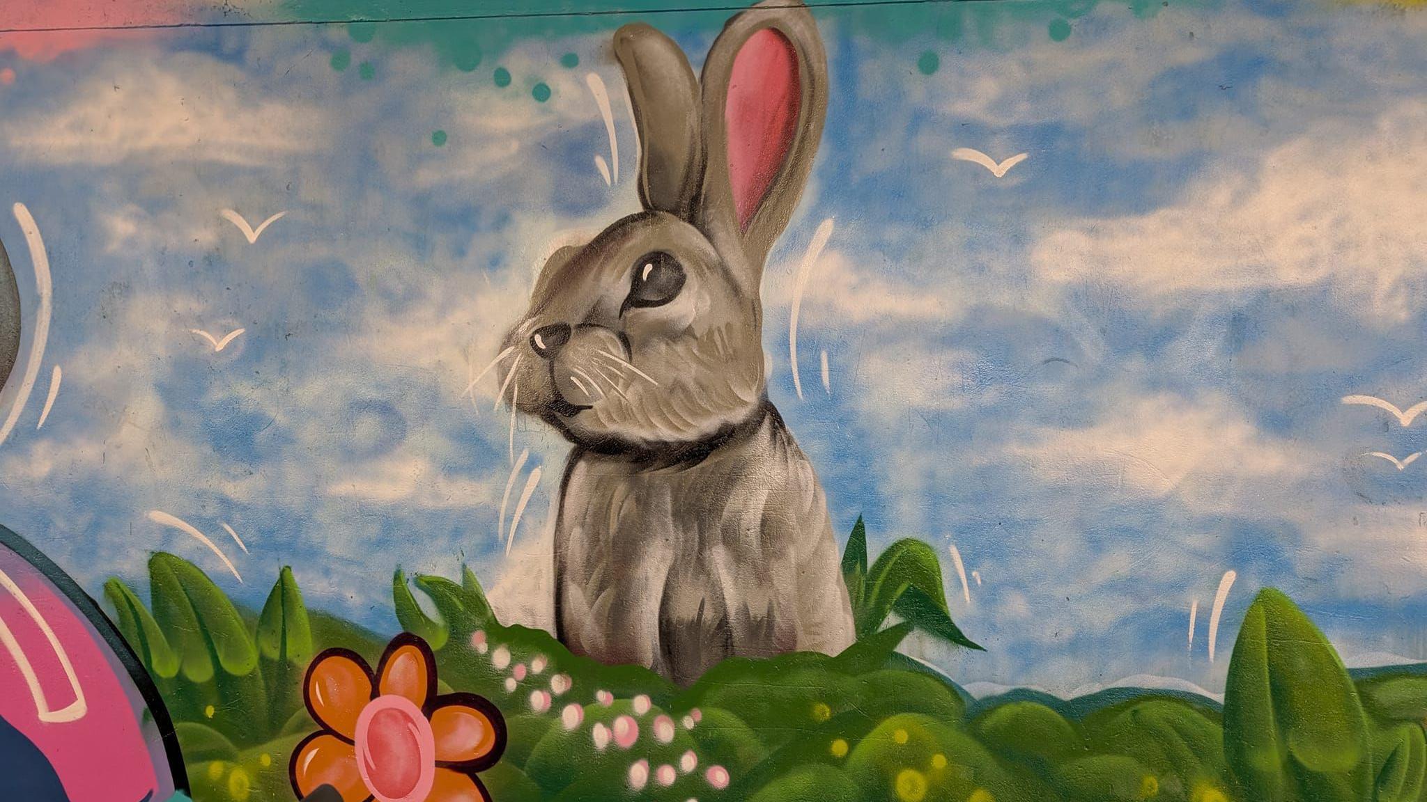 A grey painted rabbit on a blue sky background, with green grass and orange flowers underneath