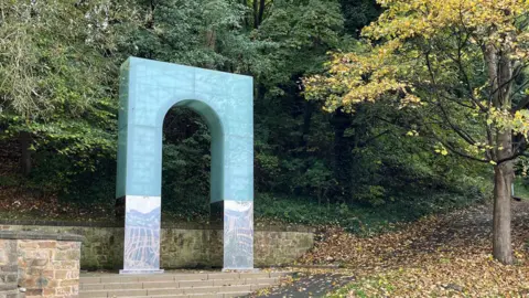 A 20ft (6m) high glass and stainless steel arch at the Pipewellgate entrance to the Riverside Park. It is placed at the top of some stone steps. It is surrounded by trees and a path leading into the main park area.