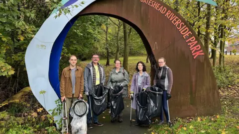 BBC Stewart Turnbull (second left) with four volunteers at an entrance to Gateshead Riverside Park. They are each carrying litter pickers and rubbish bags. They are standing in front of a large sign for the park. Behind them are trees and a path.