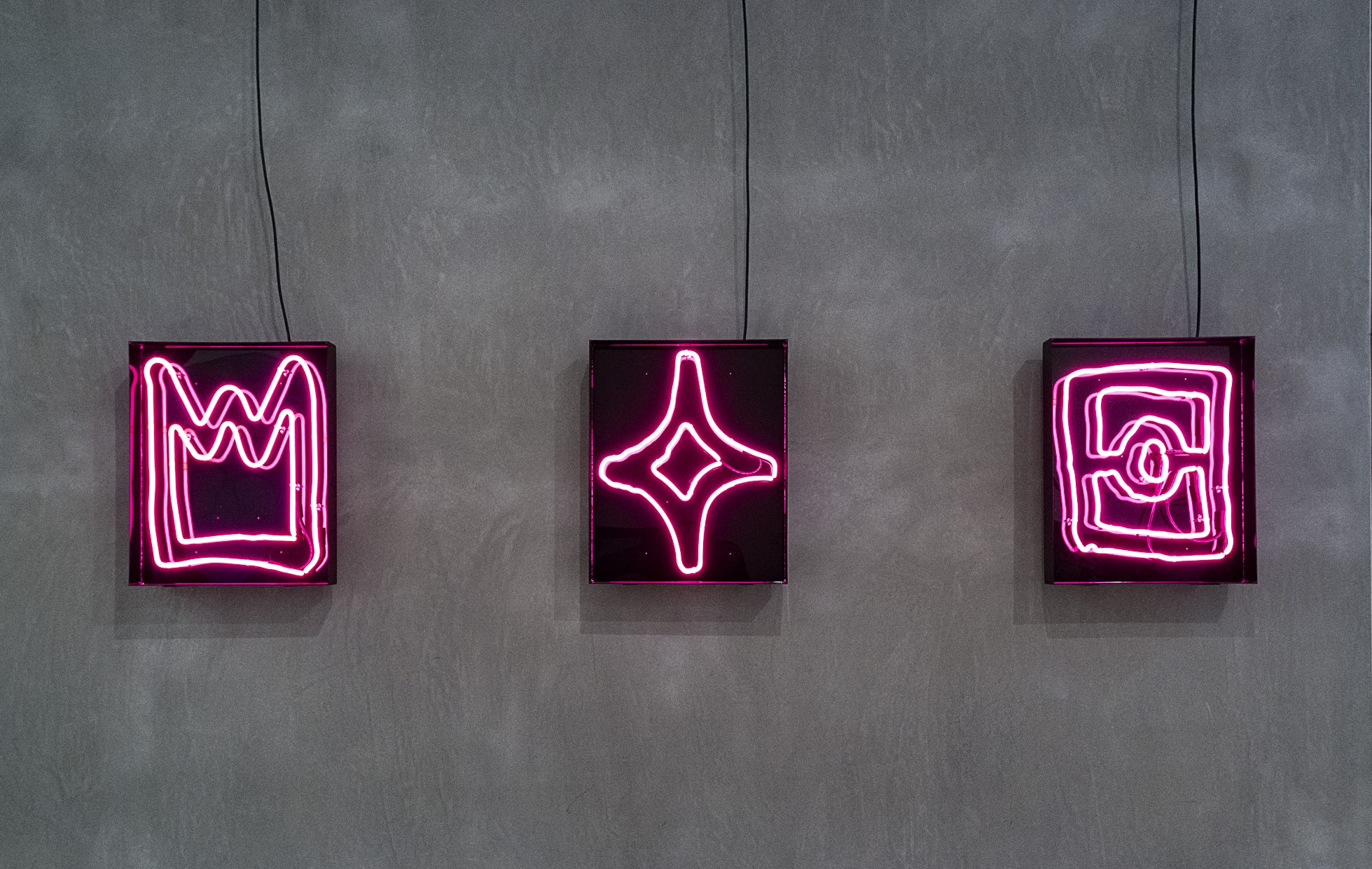 A grey wall hung with three neon pink shapes on black backgrounds: crown, diamond and flag