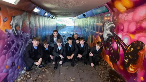 Newcastle City Council Five pupils from Studio West school, wearing their uniforms, in one of the underpasses along with artist Mark Shields and fellow artist Toby Heaps. The underpass is decorated with murals including a DJ mixer on the left-hand side of the image and a trumpet on the other.