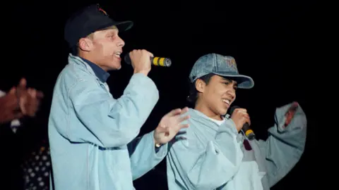 Getty Images Two rappers, wearing grey, performing a tune to a crowd of fans.