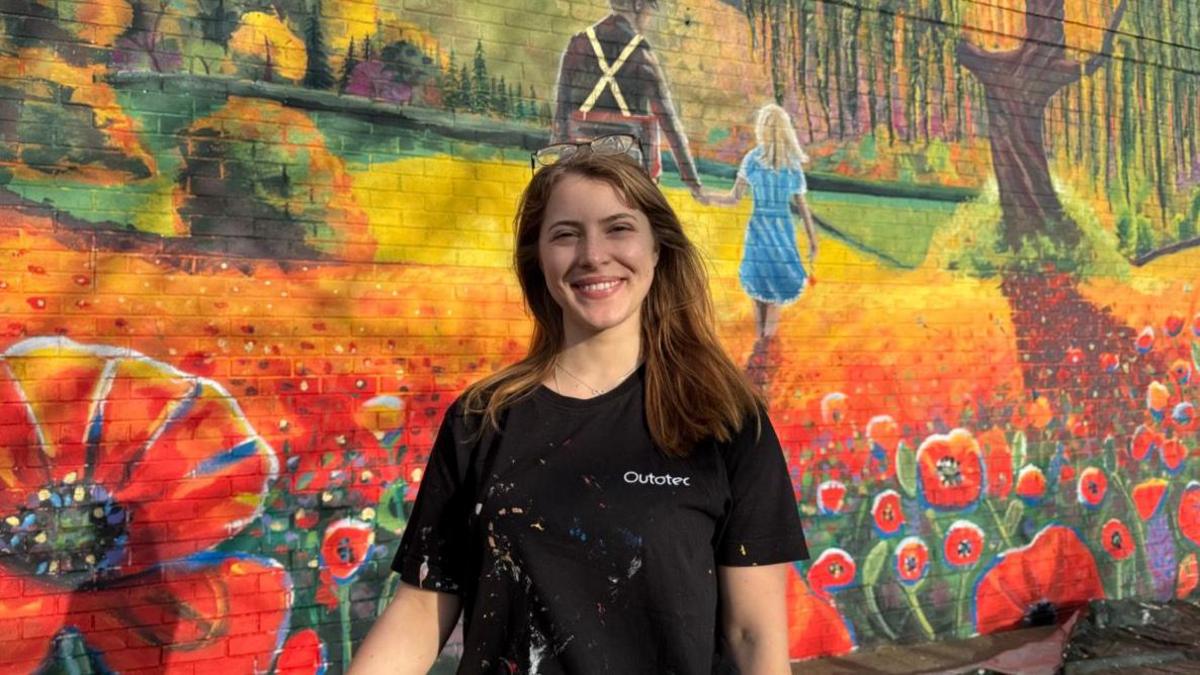 Nikita stands in front of the mural smiling. She is wearing a black t-shirt and has glasses up on the top of her head.