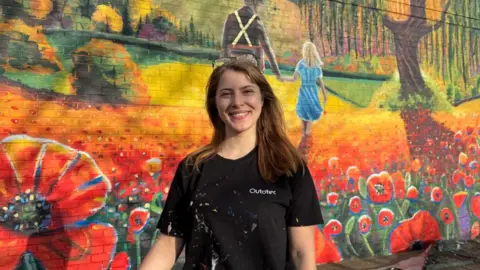 Dale Baxter/BBC Nikita stands in front of the mural smiling. She is wearing a black t-shirt and has glasses up on the top of her head.