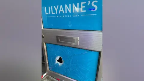 LilyAnne's Wellbeing  Blue door saying Lilyanne's Wellbeing cafe with a big hole in the glass on the bottom half there are cracks.