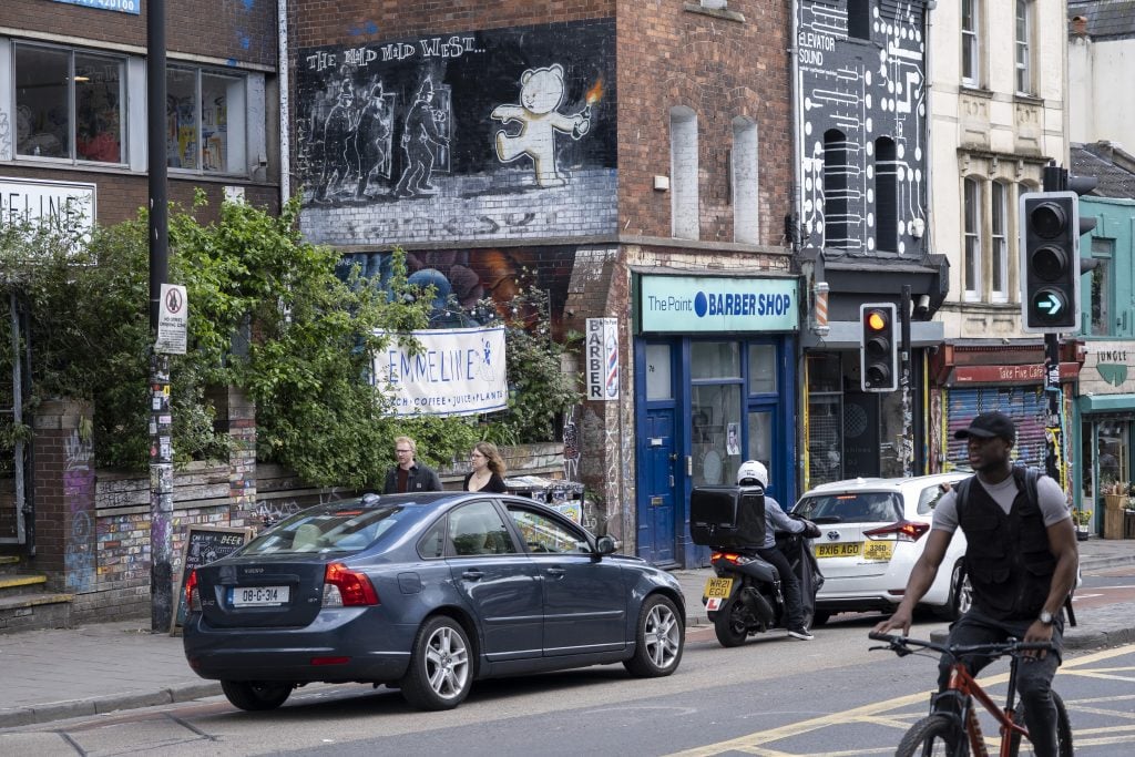 A busy street with cars traveling on a road in front of a building with a Banksy mural on its side.