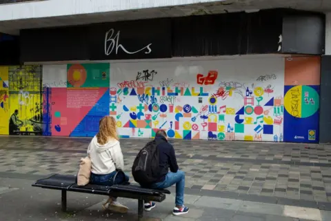 Getty Images A man and woman sit on a bench in front of a former BHS store. The windows of the store have been covered with artwork, but it has been defaced by graffiti. There is litter on the pavement.