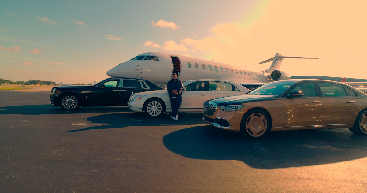 DJ Khaled stands in front of three luxury cars and a jet lined up on a runway.