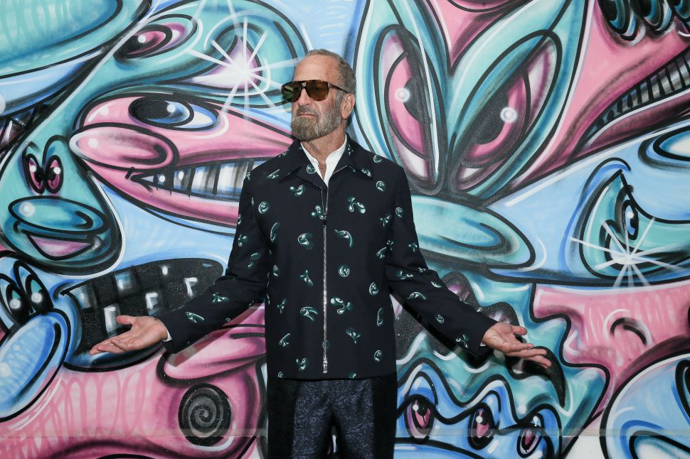 The photo shows a man with a gray beard and sunglasses standing in front of a colorful mural featuring abstract, cartoon-like faces and shapes in shades of blue, pink, and green. He is wearing a dark jacket with a pattern of small green designs, holding his hands out slightly to the sides, with a neutral expression on his face.