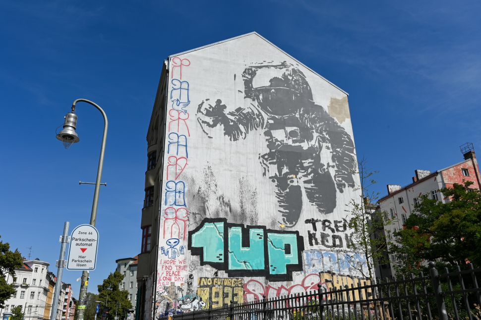 A mural on the side of a building in Berlin depicts a black-and-white astronaut holding up a peace sign. The artwork is surrounded by various graffiti, including large, colorful tags near the bottom.