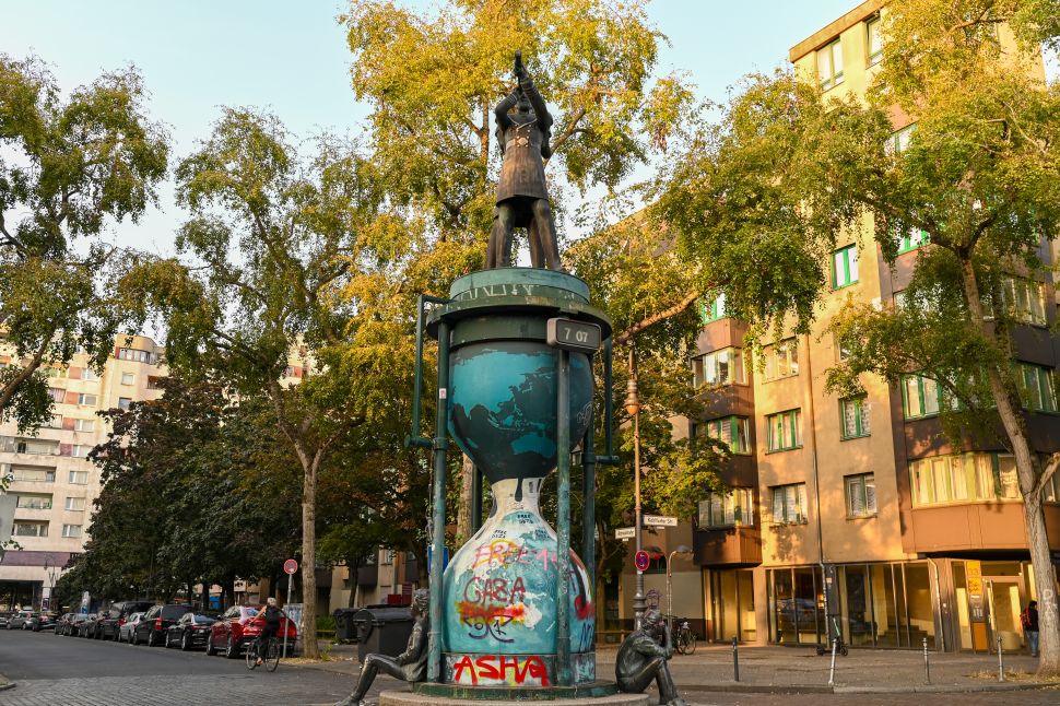 The Clessidra statue in Berlin features an hourglass-shaped base covered in graffiti with a statue of a figure standing on top, set on a street corner with trees and apartment buildings in the background.