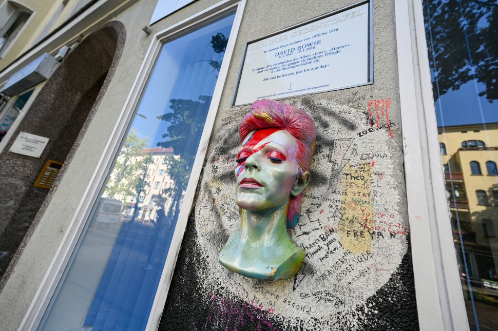 A painted bust of David Bowie as Ziggy Stardust is mounted on a wall in Berlin, below a plaque commemorating his time living in the city, with colorful graffiti and messages surrounding the artwork.