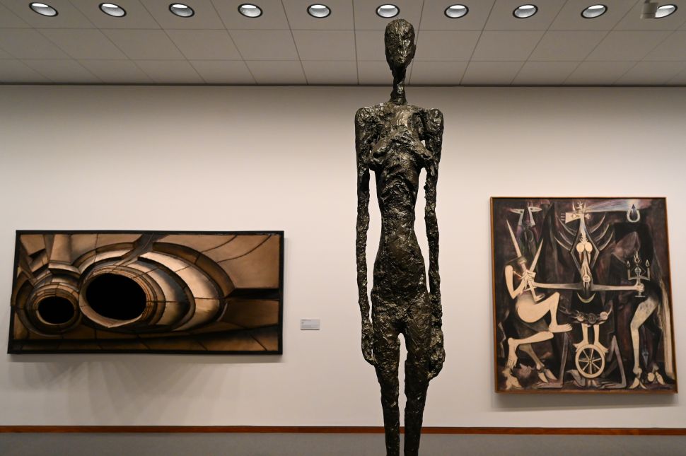 Inside the Neue Nationalgalerie, a tall, slender bronze sculpture stands in front of two abstract paintings with surreal and geometric designs, set against a plain white wall with ceiling lights above.