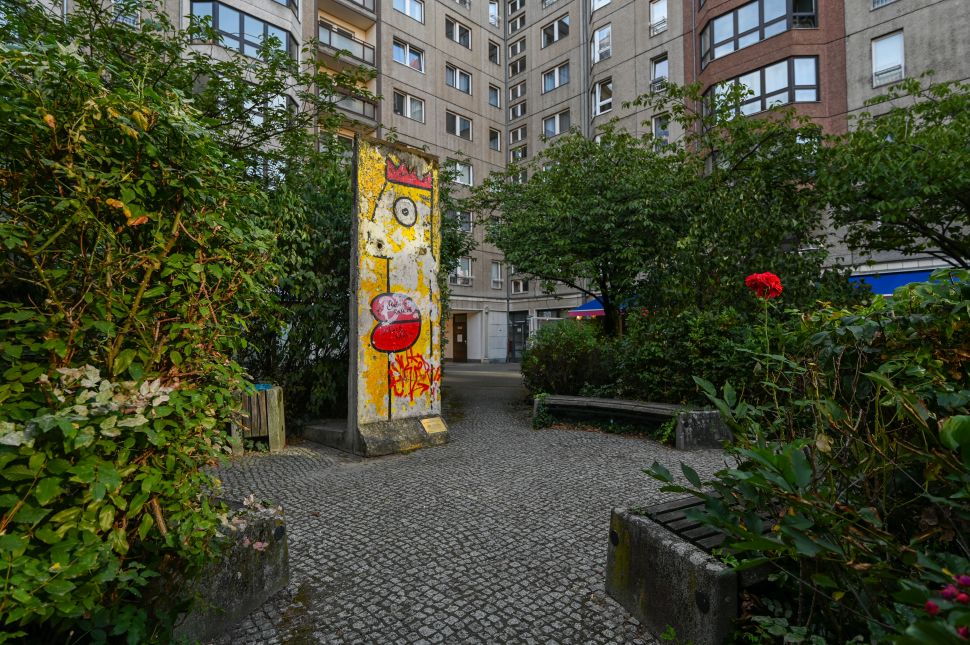 A segment of the Berlin Wall stands alone in a small garden area, decorated with colorful graffiti art, including a cartoonish face with a crown, set among trees and greenery with a nearby bench.