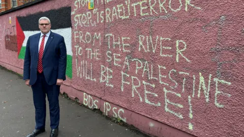 DUP DUP leader Gavin Robinson stands to the left of a wall, which has been daubed with the phrase “from the river to the sea, Palestine will be free”