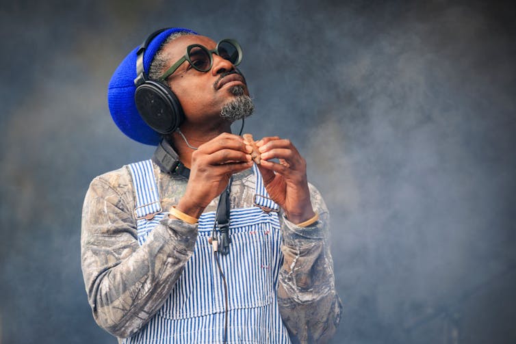 Black man wearing sunglasses, headphones and a blue beanie grasps a small flute with the fingers of both hands.