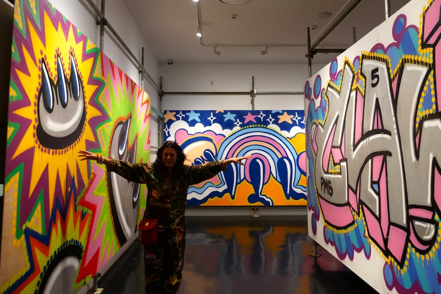 A woman stands between two vibrantly painted graffiti walls, arms outstretched wide as she leads people through the exhibit