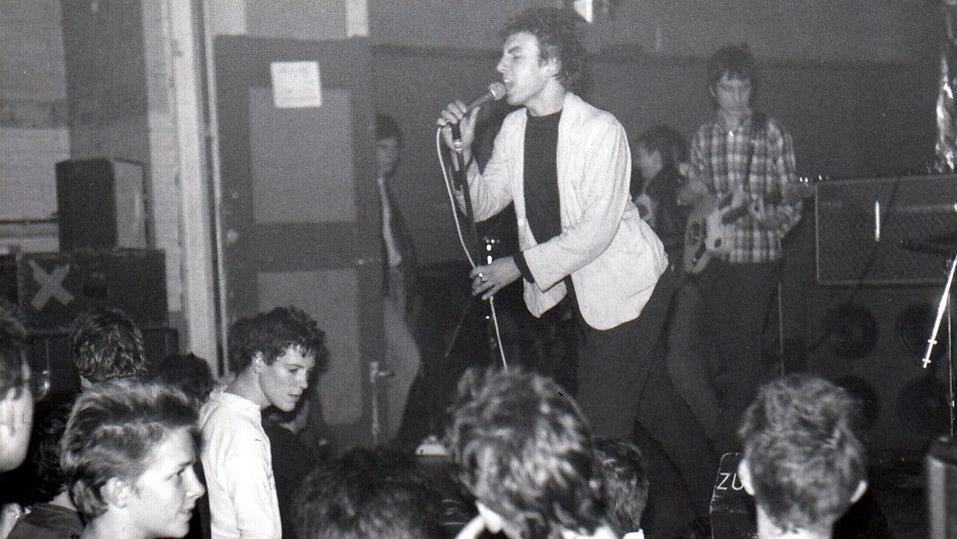 A black and white photo of a man singing into a microphone in front of a crowd of onlookers