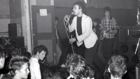 Tim Williams A black and white photo of a man singing into a microphone in front of a crowd of onlookers