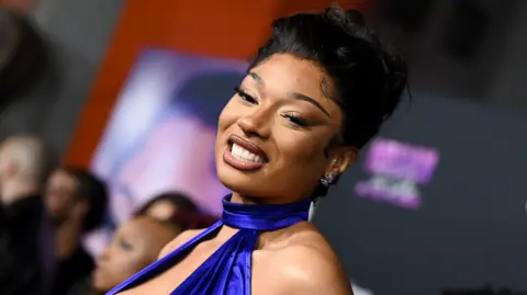 Getty Images Megan Thee Stallion pictured at the premiere of her documentary in October. Megan wears a royal blue halterneck dress and smiles at the camera. Her hair is styled in a bun and guests can be seen in the background.