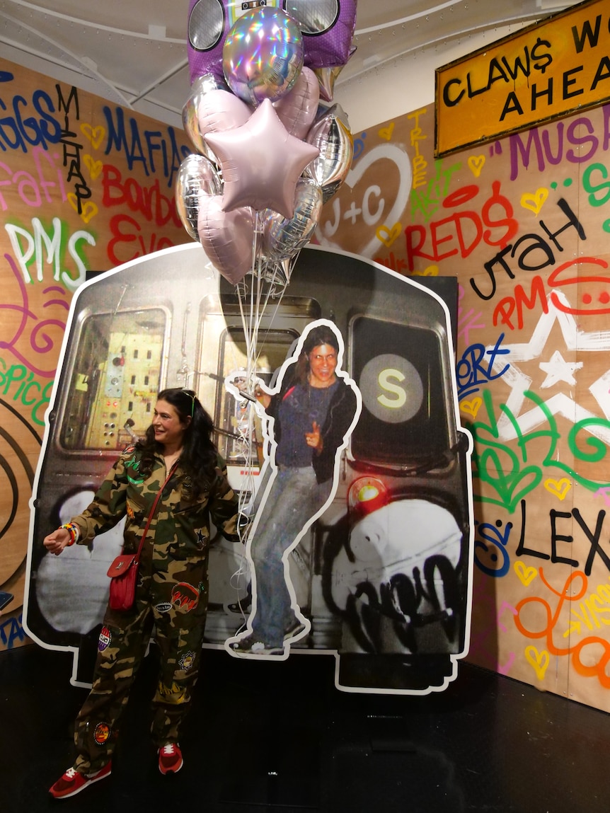 A woman stands in front of a large cardboard cutout of her younger self and a New York subway train car.