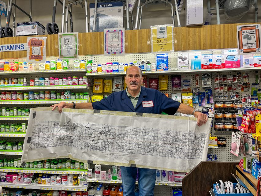 A man in a store holds a large, detailed architectural sketch. Shelves behind him display various health products, vitamins, and sale signs.