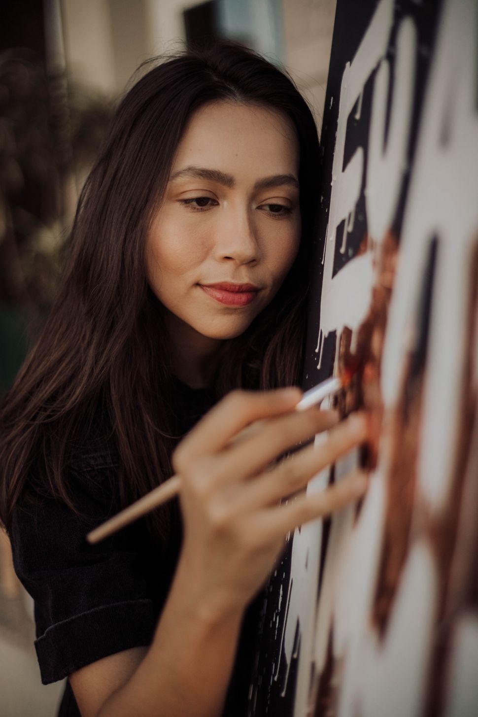 A photo of an artist painting on a canvas with her hand visible holding a brush. The artist appears focused, and her dark hair frames her face.