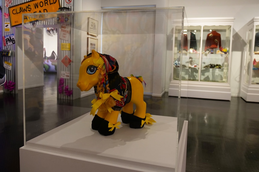 A yellow toy pony placed in a glass display box, wearing a colourful hoodie and rainbow braided mane.