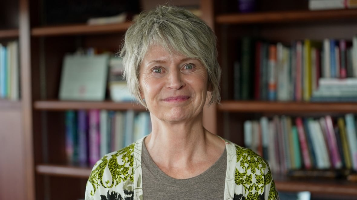 A smiling woman with short hair stands in an indoor room, a wall of bookshelves seen behind her.