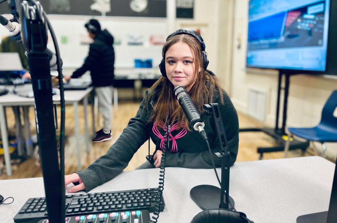 A female student wearing headhphones sits in front of a keyboard, mixing controls and a professional microphone in a classroom. Another student works in the background.