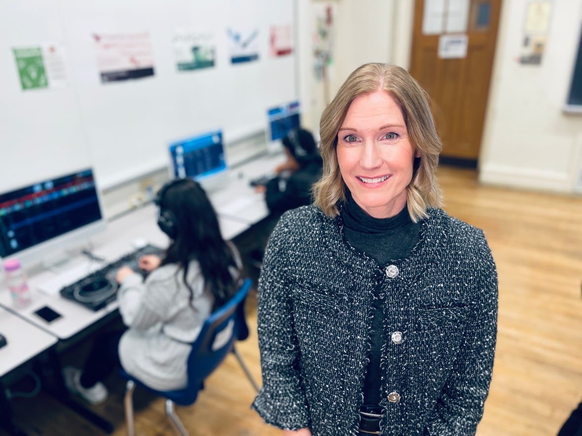 A smiling woman stands in a classroom, with students seated at computers working on music production software behind her.