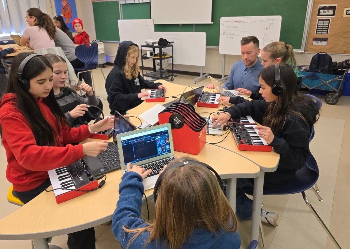 Headphone-clad elementary students gather around a low table to work on laptops and MIDI keyboard controllers in a classroom as their teacher kneels alongside a student.