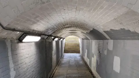 Jane Secker a dimly-lit, dirty-looking subway with steps leading out into the light outside. Graffiti is scrawled on the side one of three wall light strips. There are patches of white paint over the grey tiles on the walls. The ceiling is dirty and curved like in a tunnel.