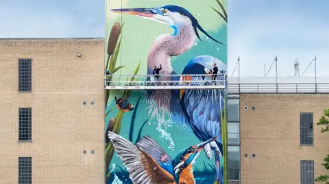 Street Art Atlas Peachzz and her assistant on a platform, pulled up the side of the building by a crane. They are working on the painting described above, with her holding up a spray paint can. They are about two thirds of the way up the building. Most of the painting is complete, but she is adding texture.