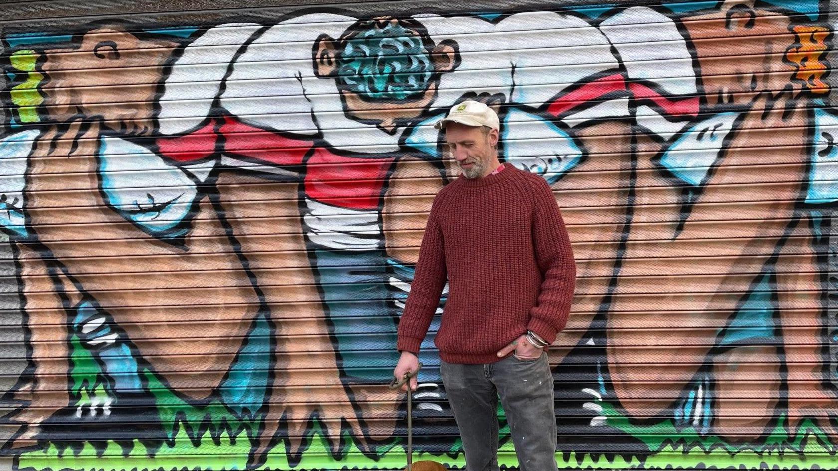 Mural depicting rugby players on the shutters of an empty shop. The artist is in black jeans and a red jumper. He has a grey beard and white cap