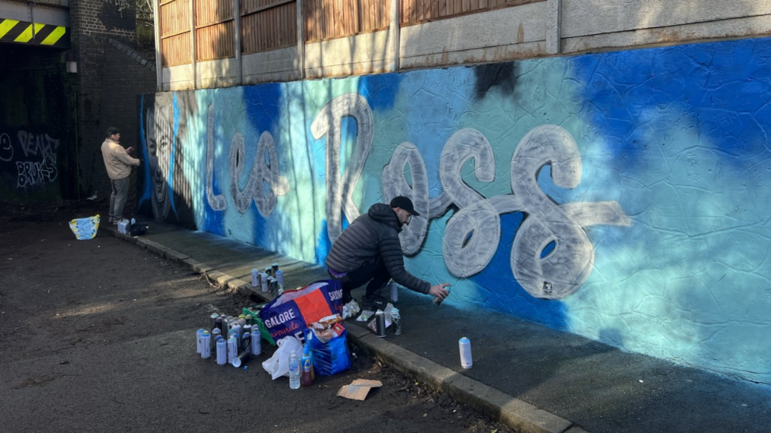 Two men stand near the mural, painted on a wall near a railway bridge. The blue mural has the words Leo Ross painted on in white lettering.