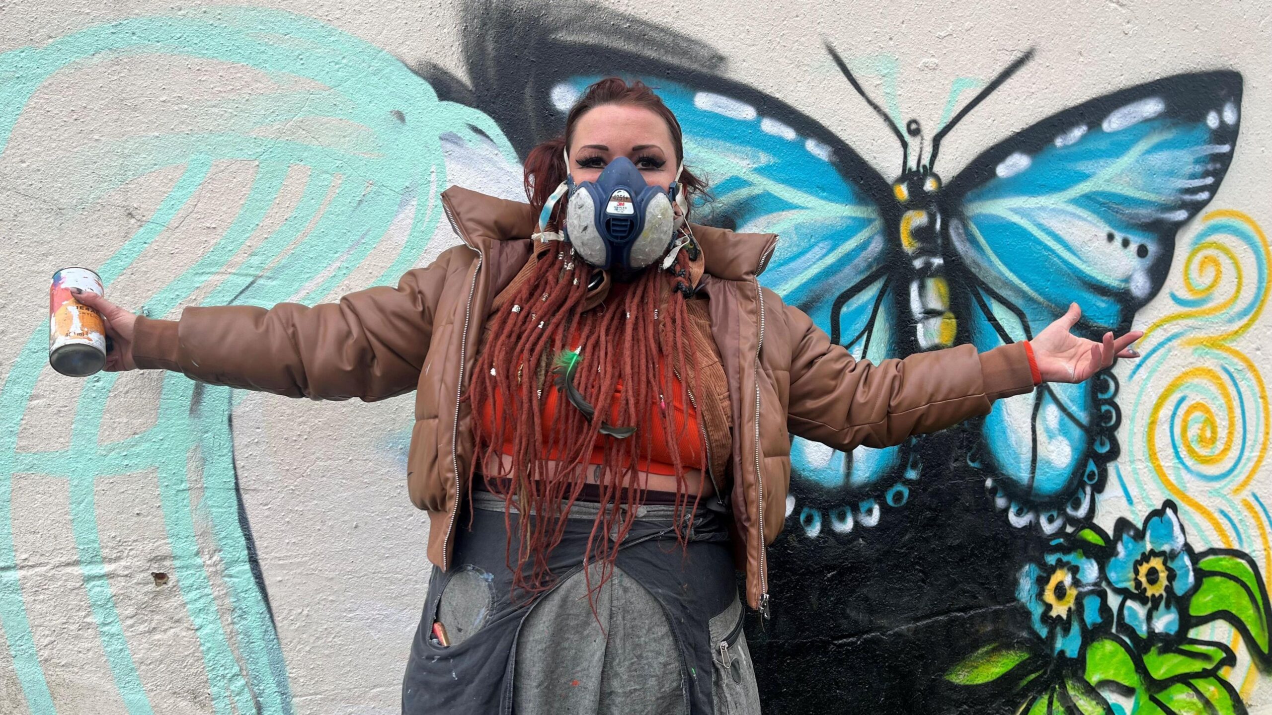 Sarah Marshall, a woman who is wearing a red t-shirt and brown jacket, is stood with her arms open whilst posing for the camera with a spray can. Behind her is a partially-completed mural which features a butterfly and flowers.