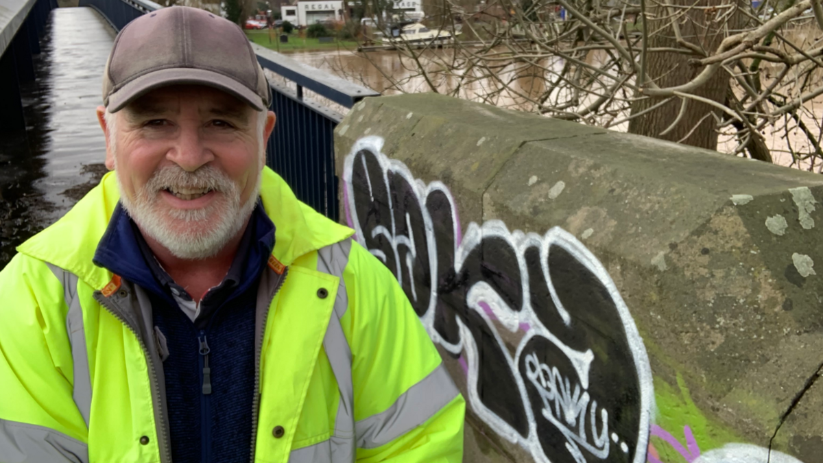 A man with a white beard is wearing a hi-vis jacket and a dark baseball cap. He is standing by a bridge which has been tagged with graffiti.