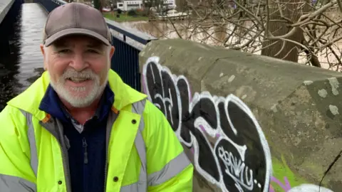 LDRS A man with a white beard is wearing a hi-vis jacket and a dark baseball cap. He is standing by a bridge which has been tagged with graffiti.