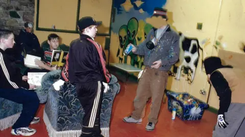 Peter de Boer Banksy addresses a room of young people, holding a blue spray can and has a mask hanging from his neck. He is wearing a bucket hat, a paint-covered zip-up top and brown trousers. Behind them is a half-painted mural featuring aliens, spaceships and a cow.