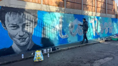 BBC A man touches a wall on which a gigantic blue mural has been painted. On the left is Leo Ross's face, neck, and shoulders. On the right is his name, painted on the wall in white writing.