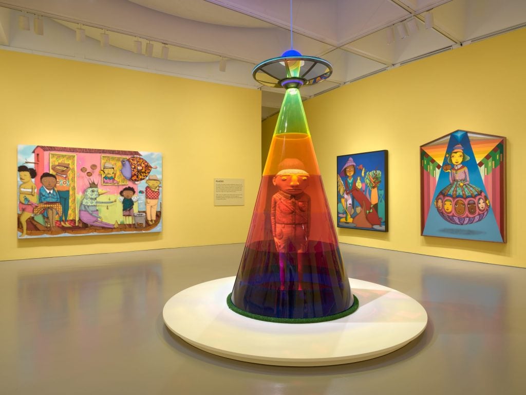 exhibition room inside a museum with several large colorful paintings attached to a big yellow wall with a cone-shaped tree in the center on a round pedestal