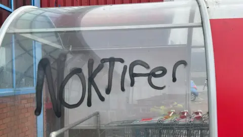 Great Yarmouth Borough Council A glass trolley bay with the word 'Noktifer' spray painted black on the side. Trolleys with red handles are located within the trolley bay.