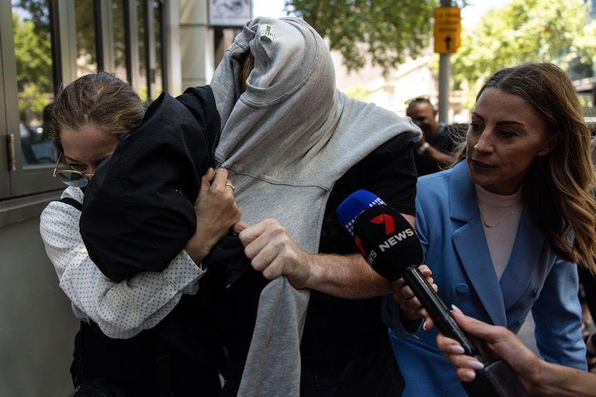 Matthew Raoul White has a jumper draped over his face between a supporter and a journalist as he leaves court.